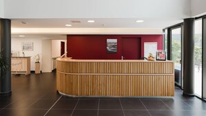 A reception area with a wooden reception desk, a red accent wall, and a view through large windows. - Quality Aparthotel Saint-Malo (Saint-Malo)