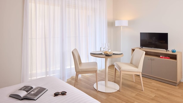 A hotel room with a bed, a round table with chairs, a TV on a wooden cabinet, and a large window with sheer curtains. - Quality Aparthotel Voiron (Voiron)