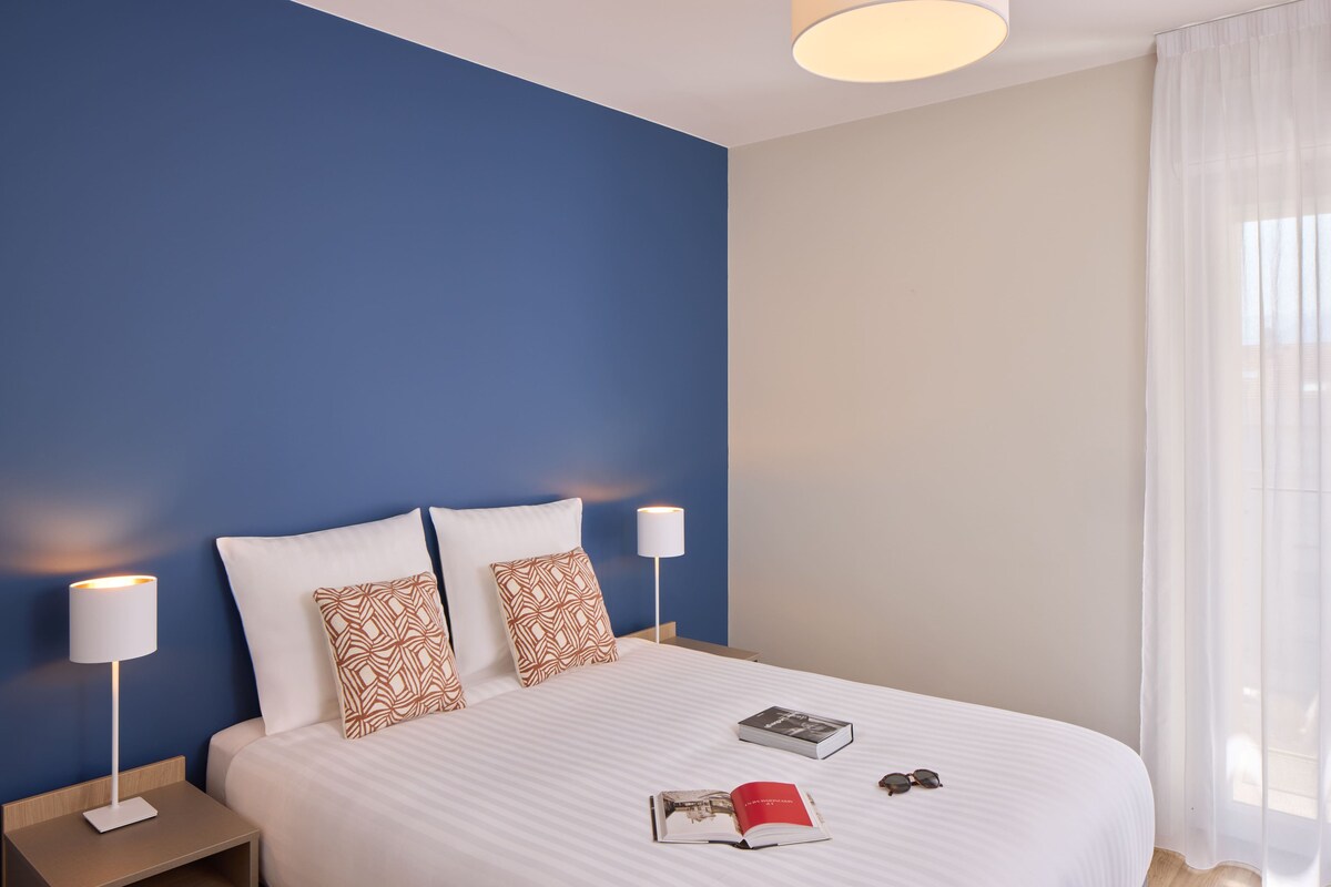 A neatly made bed with two pillows, a bedside lamp, and a book on the bed.