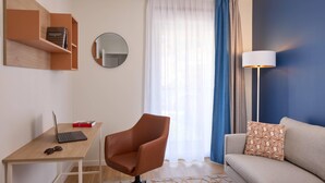Ένα μοντέρνο σαλόνι με ένα ξύλινο γραφείο, μια καφέ δερμάτινη πολυθρόνα, έναν καναπέ με διακοσμητικά μαξιλάρια, έναν στρογγυλό καθρέφτη και ένα μεγάλο παράθυρο με λεπτά, διάφανα κουρτίνια.