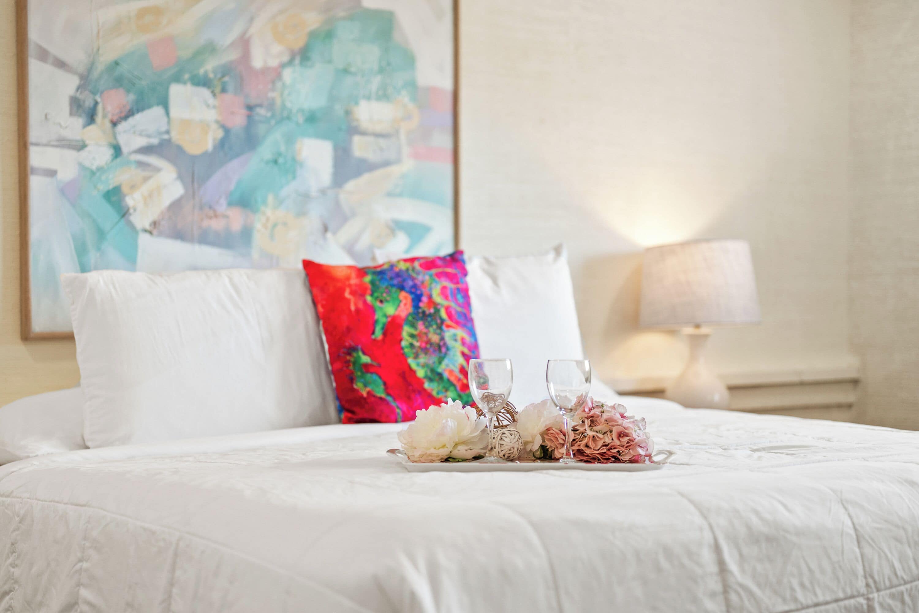 A neatly made bed with white linens, a colorful pillow, and a tray with flowers and glasses.