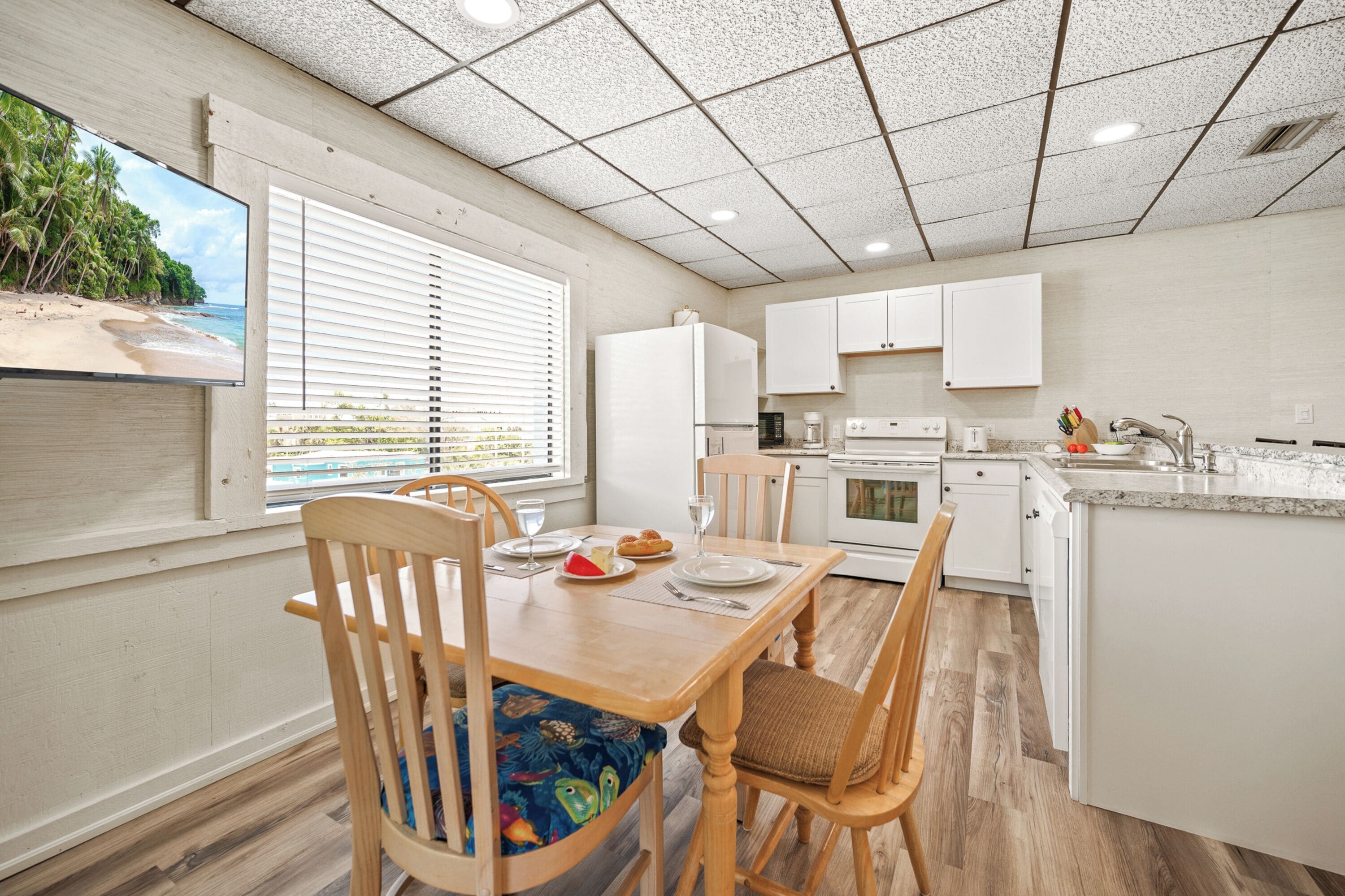 Una cocina con armarios blancos, una mesa de comedor con sillas, un televisor montado en la pared y una ventana con persianas.