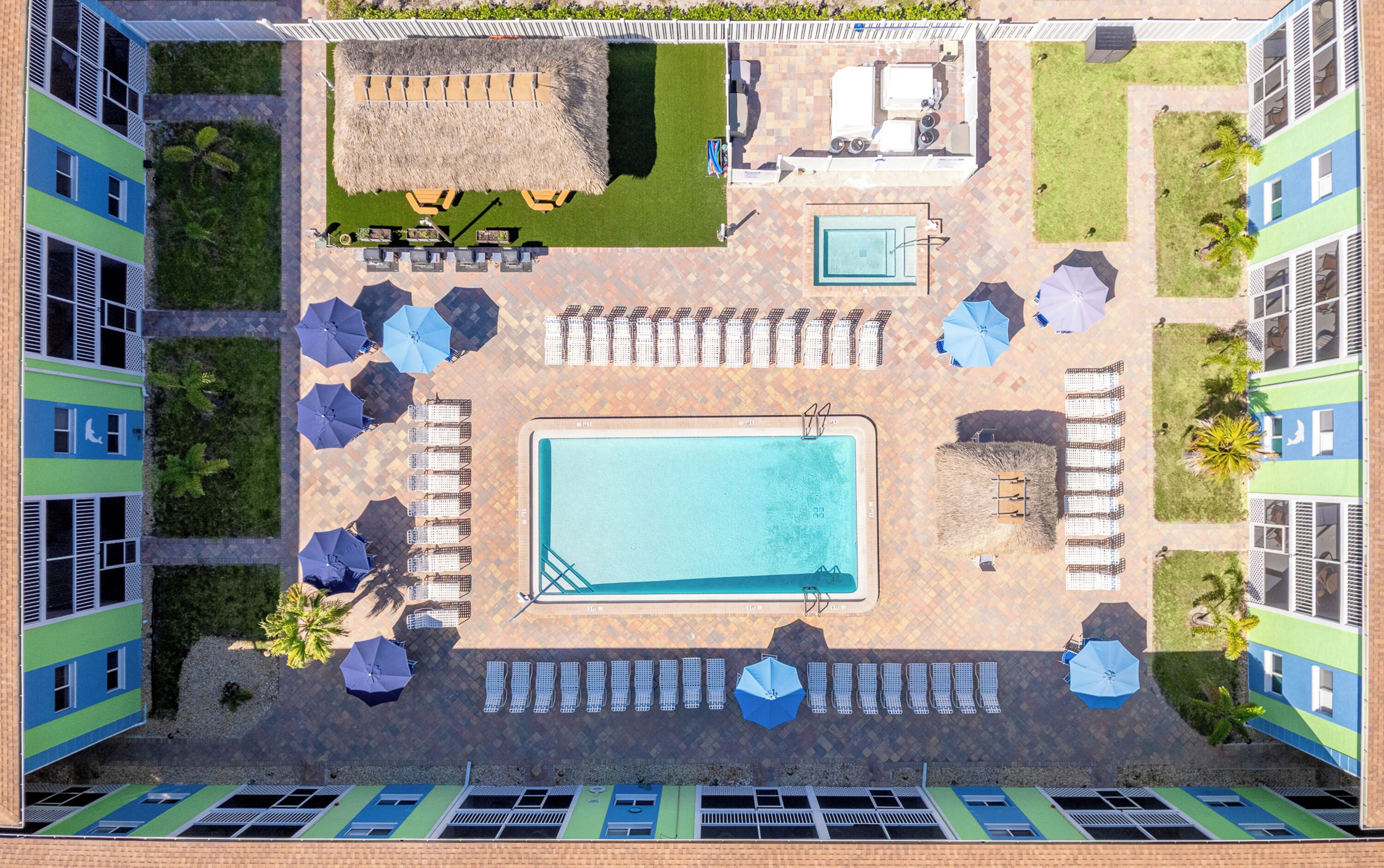 An aerial view of a hotel with a central swimming pool, lounge chairs, umbrellas, and a small garden area.