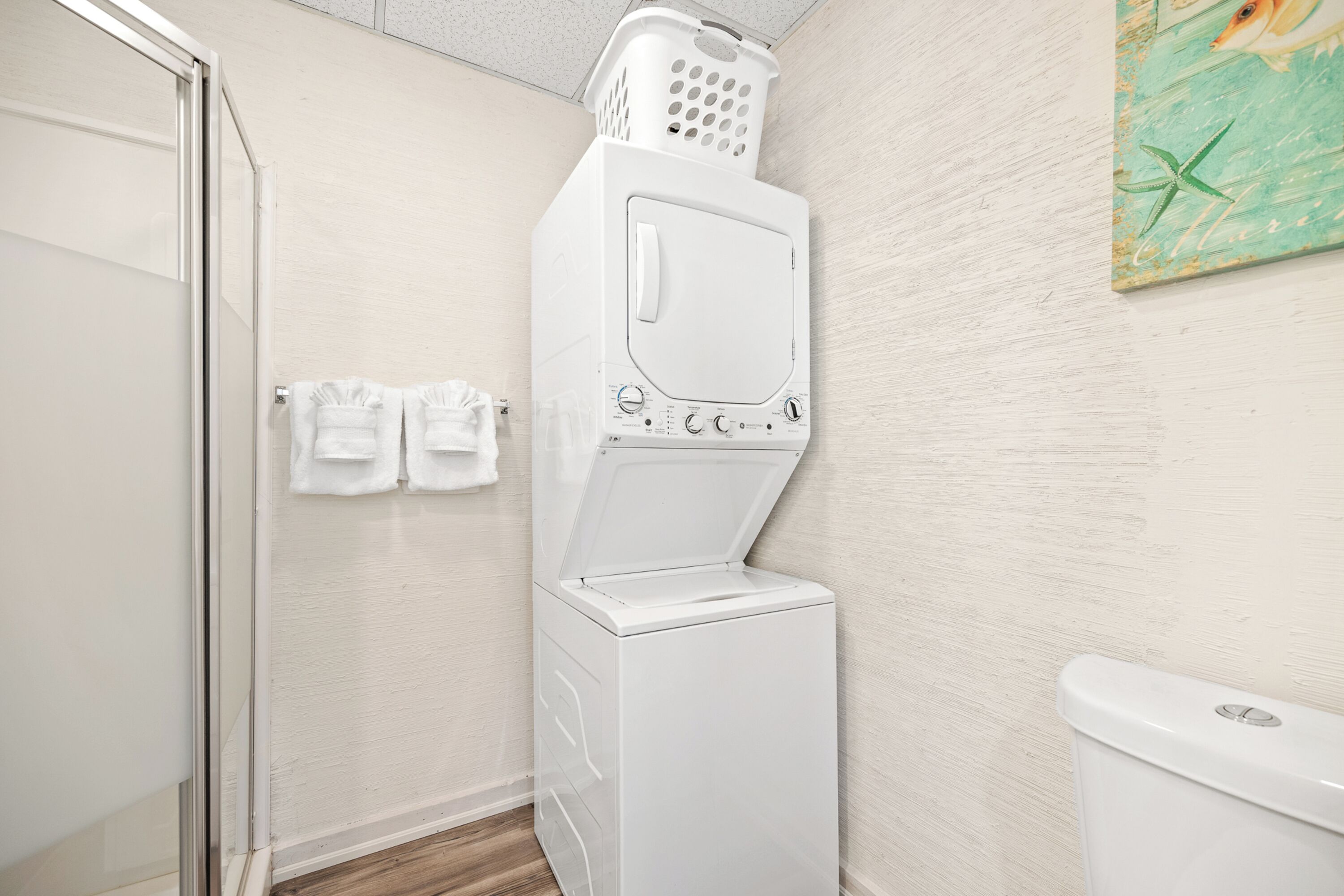 A compact laundry room with a front-loading washer and dryer, a glass shower door, and a wall-mounted towel rack.