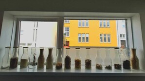 Una finestra che si affaccia su un edificio giallo con numerose finestre, con diversi vasi di vetro contenenti vari materiali.