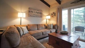 Un salon chaleureux avec un canapé d’angle, deux lampadaires, une table basse en bois et une grande fenêtre donnant sur les arbres.