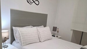 Cama bem arrumada com lençóis brancos, cabeceira cinza e decoração em formato de coração na parede.