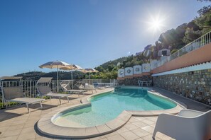 Zona de piscina con sillas de descanso y sombrillas, rodeada de muros de piedra y un cielo despejado.