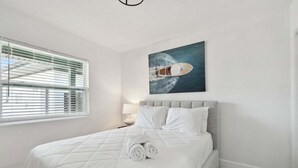 Een netjes opgemaakt bed met witte lakens en opgerolde handdoeken, een grijs gestoffeerd hoofdbord en een schilderij van een boot aan de muur.