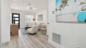 Una habitación moderna y luminosa, con piso de madera, sofá blanco, una mesa de comedor con sillas y un cuadro de una escena de playa en la pared.