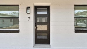 Una casa con puerta principal negra, el número 7 en la pared y dos ventanas con persianas.