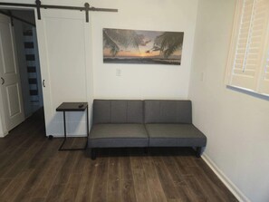Ein Zimmer mit grauem Sofa, einem kleinen Tisch, einem Sonnenuntergangsbild und Holzboden.