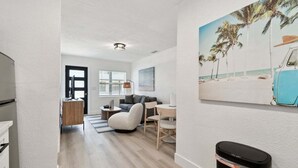 Uma sala de estar moderna com um sofá, uma mesa de jantar e uma pintura de uma paisagem praiana.