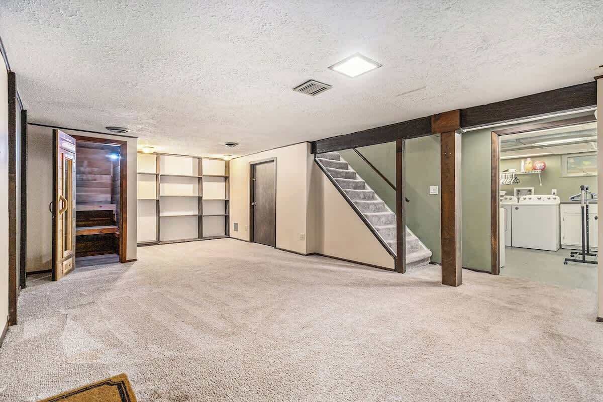 Una habitación amplia con piso alfombrado, una escalera y una cocina visible a través de una puerta abierta.
