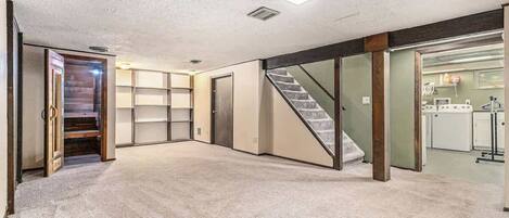 Una habitación amplia con piso alfombrado, una escalera y una cocina visible a través de una puerta abierta.