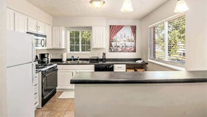 Una cocina moderna con armarios blancos, encimera negra y una isla grande. Hay dos luces colgantes, una ventana con persianas y un cuadro en la pared.