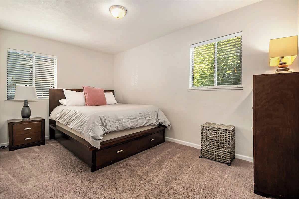 A bedroom with a bed, nightstands, a lamp, a window with blinds, and a small wicker basket.