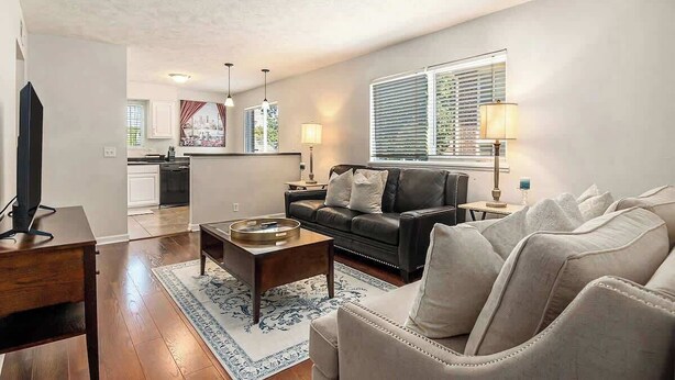 Una sala de estar con sofá, mesa de centro y televisor sobre un mueble bajo. Se observa una cocina al fondo, con armarios y una ventana.