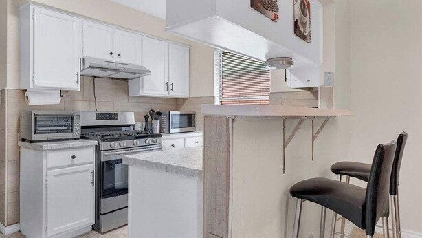A modern kitchen with white cabinets, a built-in oven, a microwave, and a gas stove. There are two black bar stools and a window with blinds. - Modern 5BR Home |Cozy Fireplace, Office & Downtown (Austin)