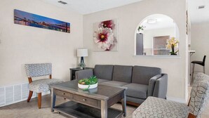 A living room with a grey sofa, patterned chairs, a coffee table with a plant, a painting of a cityscape, and a mirror reflecting the room. - Modern 5BR Home |Cozy Fireplace, Office & Downtown (Austin)