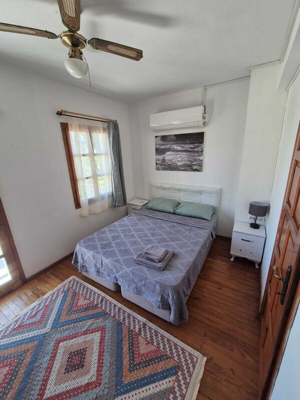 A bedroom with a bed, a ceiling fan, a window with curtains, a wall-mounted air conditioner, a bedside table with a lamp, and a patterned rug.