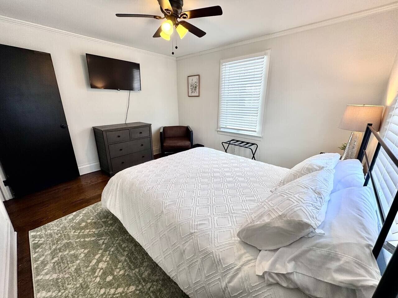 Een slaapkamer met een bed, een plafondventilator, een televisie, een dressoir, een stoel en een raam met jaloezieën.