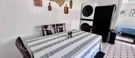 Un comedor con una mesa dispuesta para cuatro, sillas, una lavadora y una cama al fondo.