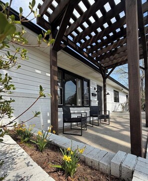 Une terrasse couverte agrémentée d’une pergola en bois, un bâtiment blanc avec le numéro « 1231 » sur la porte, et un espace détente avec deux chaises.