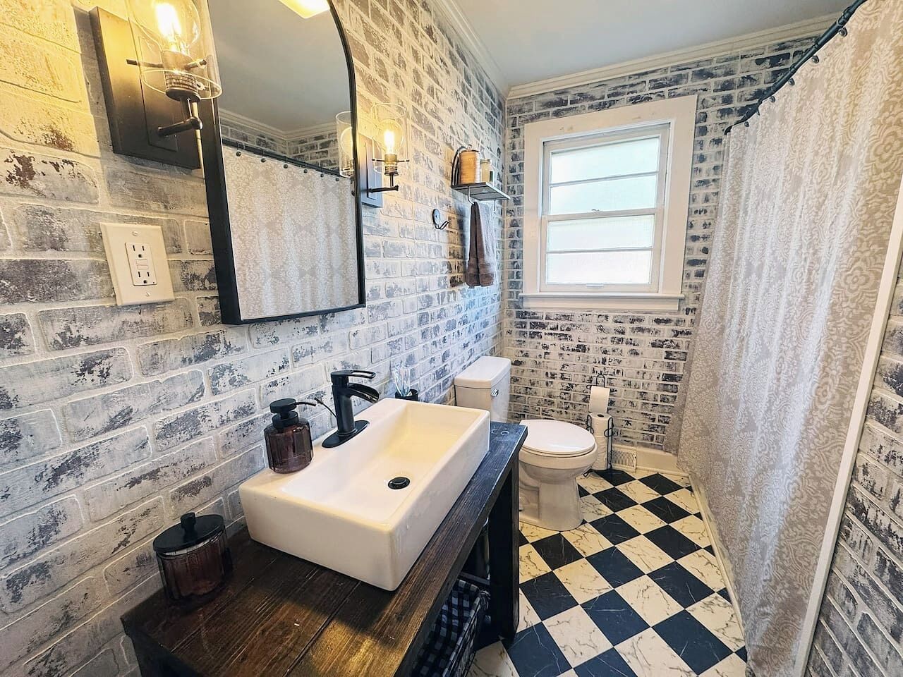 Un bagno con un lavandino bianco, pavimento a scacchi bianchi e neri, una finestra e un muro in mattoni.