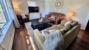 A living room with a sofa, coffee table, TV, and ceiling fan. - Hidden Getaway | Fire Pit | 1K,2Q/1 bath (Eden)