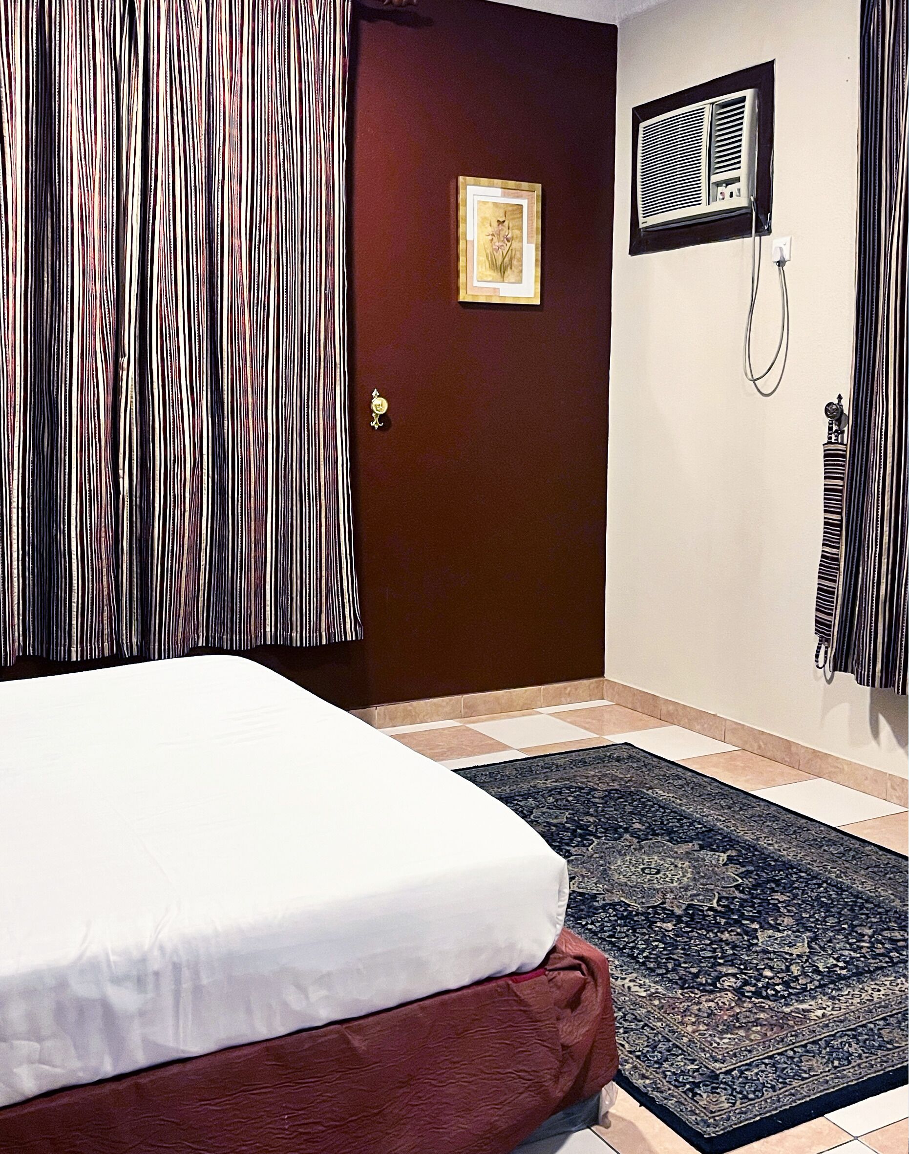 A hotel room with a bed, a dark-colored rug, a door with a picture, and a wall-mounted air conditioning unit.