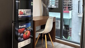 A compact living space with a black refrigerator, a wooden desk, a white chair, and a large window with curtains. - NQ Apartment (Hanoi)