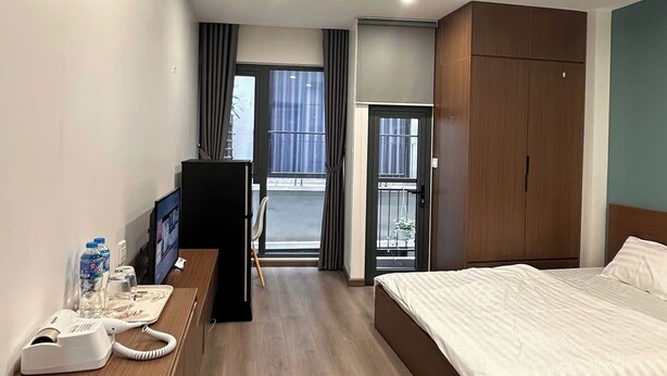 A hotel room with a bed, a desk with a telephone, an air conditioner, and a view of the outside through a window. - NQ Apartment (Hanoi)