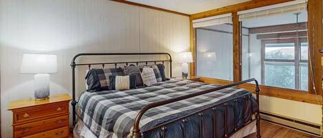 A bedroom with a metal bed frame, a wooden nightstand with drawers, a large window with blinds, and a view of the outdoors.