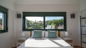 Een slaapkamer met een groot raam, een bed met witte beddengoed en groene kussens, twee nachtkastjes met lampen, en uitzicht op een groen landschap.