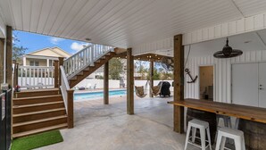 A covered outdoor area with a wooden staircase leading to a pool, a bar area with stools, and a hanging fan.