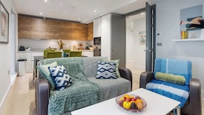 Een moderne woonkamer met een bank, een eettafel met fruit, en een keuken op de achtergrond.