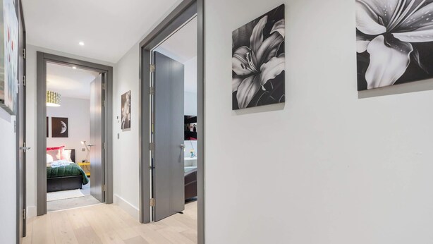 Een gang met een bed op de achtergrond, een deur naar een andere kamer, en twee bloemschilderijen aan de muur.