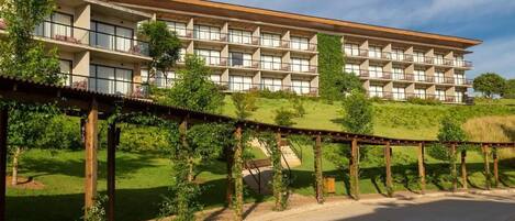 A multi-story hotel with a flat roof and balconies, surrounded by greenery and a paved walkway.