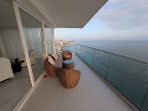 Un balcón con mobiliario de mimbre, puerta de cristal y vistas al mar y al paisaje urbano.
