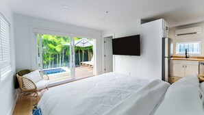 Quarto de hotel moderno com uma cama grande, televisão na parede, porta de vidro deslizante para área de piscina e cozinha compacta.