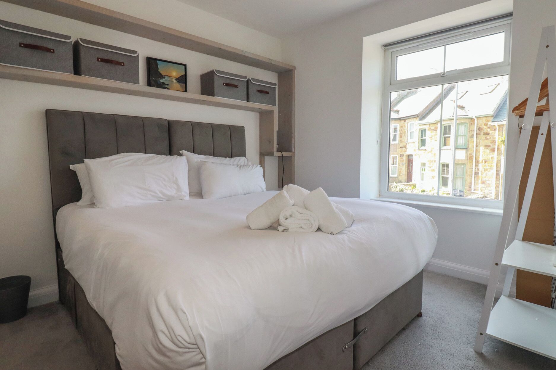 Een net opgemaakt bed met kussens en een opgerolde handdoek, een raam met uitzicht op gebouwen, en opbergkisten op een plank boven het bed.