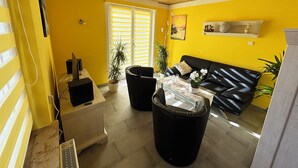 A living room with a yellow wall, a black leather sofa, a glass coffee table, and two black armchairs. There is a television on a stand, a plant, and a window with blinds.