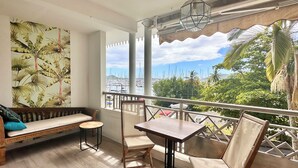 Un balcon avec un banc en bois, une petite table ronde et deux chaises. Il donne sur une marina où l’on peut admirer des bateaux et des palmiers.
