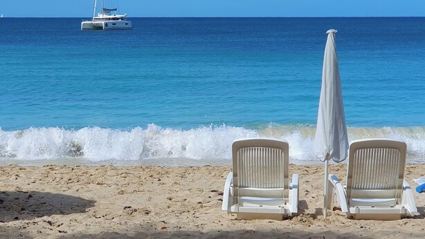 Une plage avec deux chaises longues blanches et un parasol, un voilier à l’horizon et un ciel bleu dégagé.