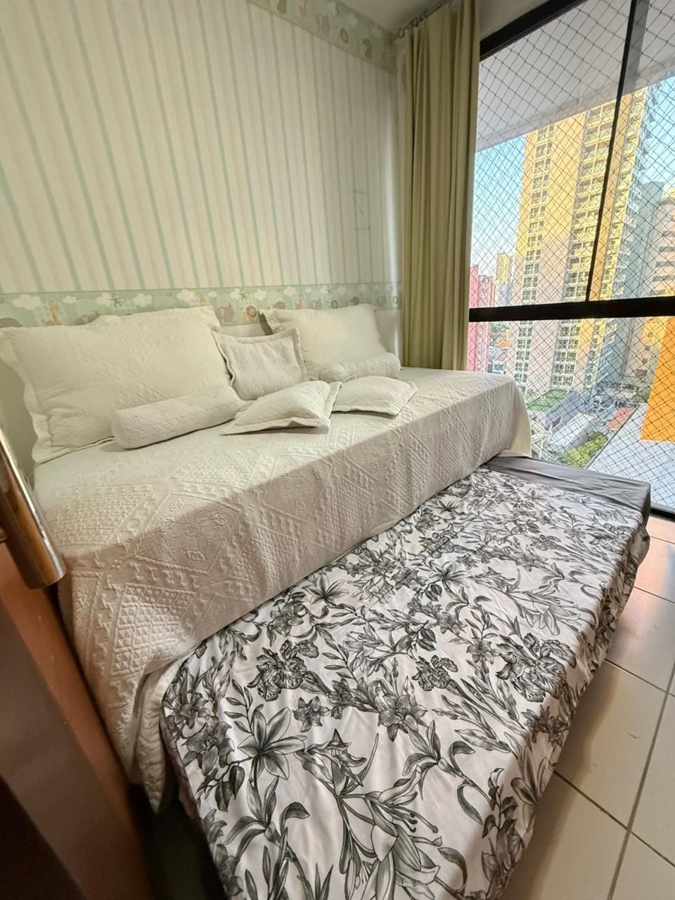 A bed with a floral bedspread and a plain headboard, a window with a view of buildings, and a curtain.