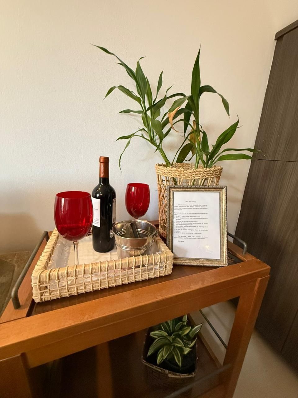 A wooden table with a woven placemat, two red wine glasses, a bottle of wine, a metal container, a framed menu, and a potted plant.