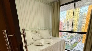 Un dormitorio con cama, cortinas, una ventana con vistas a edificios altos y suelo de baldosas.