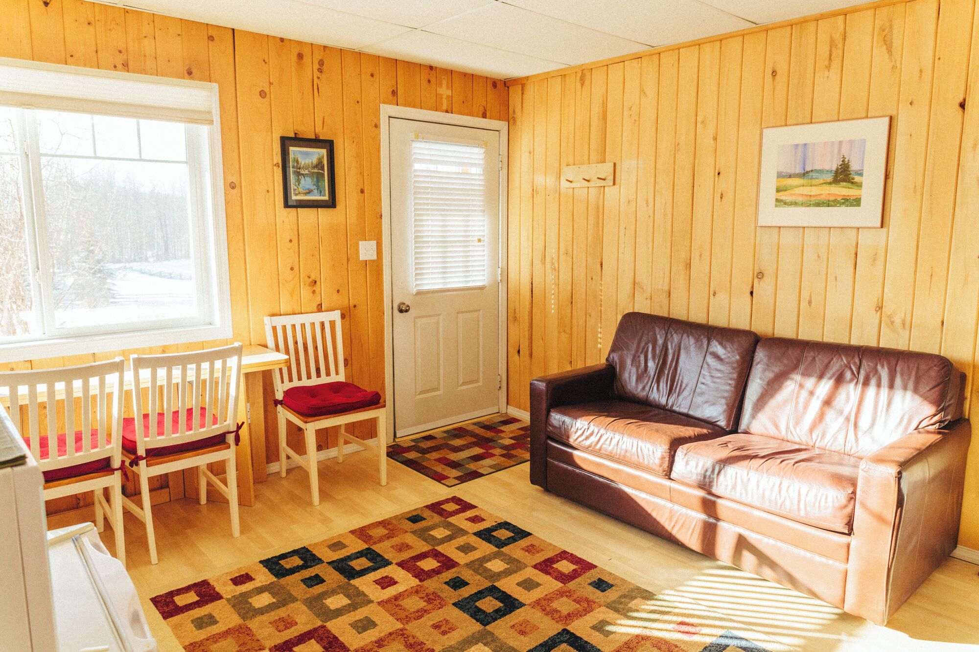 A cozy room with wooden walls, a brown leather couch, a wooden table with chairs, a patterned rug, and a door with a window.