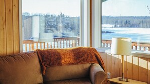 A cozy room with a wooden cabin interior, featuring a couch with a blanket, a lamp, and large windows offering a view of a snowy landscape. - Family run swiss-style chalet, sauna and ski retreat near Twin Lakes (Winfield)